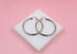A pair of shiny silver hoop earrings rests on a white square display block against a soft pink background.