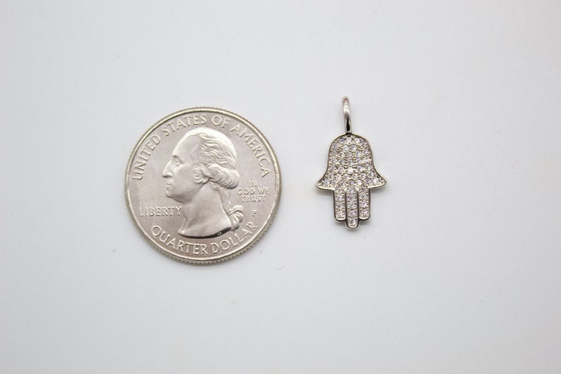A diamond-encrusted silver hamsa hand pendant rests next to a US quarter coin for scale on a white surface.