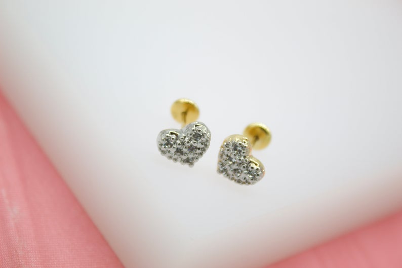 A pair of gold heart-shaped stud earrings encrusted with small sparkling diamonds sits on a white display surface.