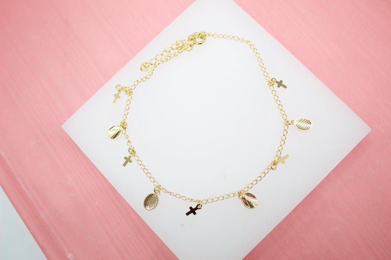A delicate gold-plated anklet featuring hanging cross and oval medal charms rests on a white display surface against a pink background.