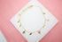 A delicate gold-plated anklet featuring hanging cross and oval medal charms rests on a white display surface against a pink background.