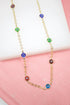 A gold chain necklace displays colorful evil eye charms in blue, green, red, and purple against a pink and white background.