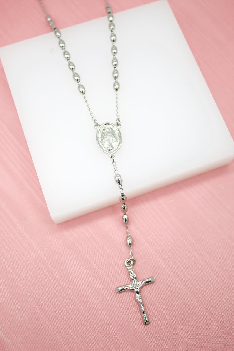 A shiny silver rosary necklace with a crucifix rests on a white display block against a pink background.