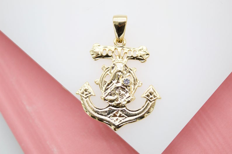 A polished gold anchor pendant featuring an intricate nautical wheel design with a central diamond-like stone lies flat on a dual-toned white and pink background.