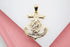 A polished gold anchor pendant featuring an intricate nautical wheel design with a central diamond-like stone lies flat on a dual-toned white and pink background.