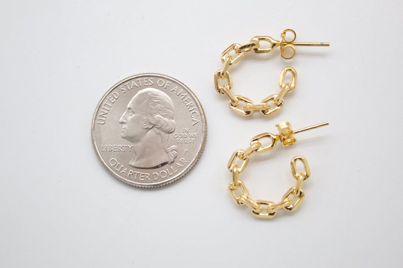 A pair of gold chain-link hoop earrings rests next to a US quarter coin on a white surface for size comparison.