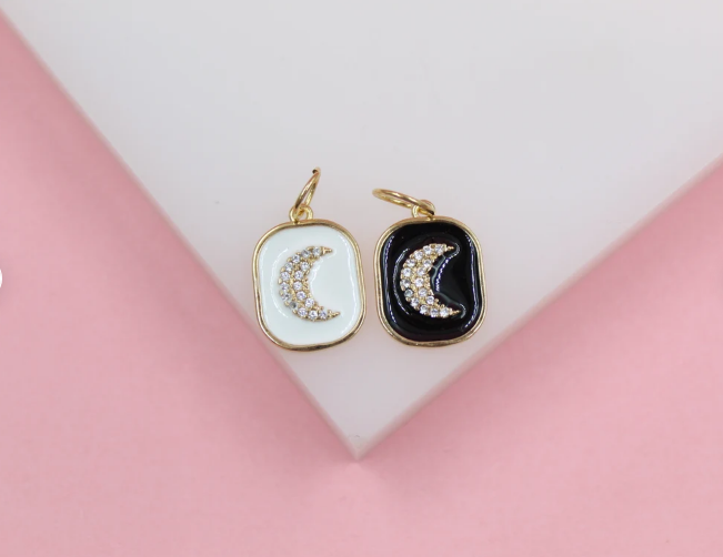 Two gold crescent moon charms with sparkling crystal pave rest side-by-side on a white display against a soft pink background.