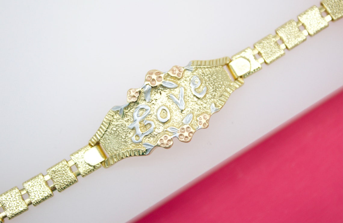 A tri-color gold ID bracelet features the word "LOVE" in raised white gold letters framed by delicate floral accents against a textured gold background.