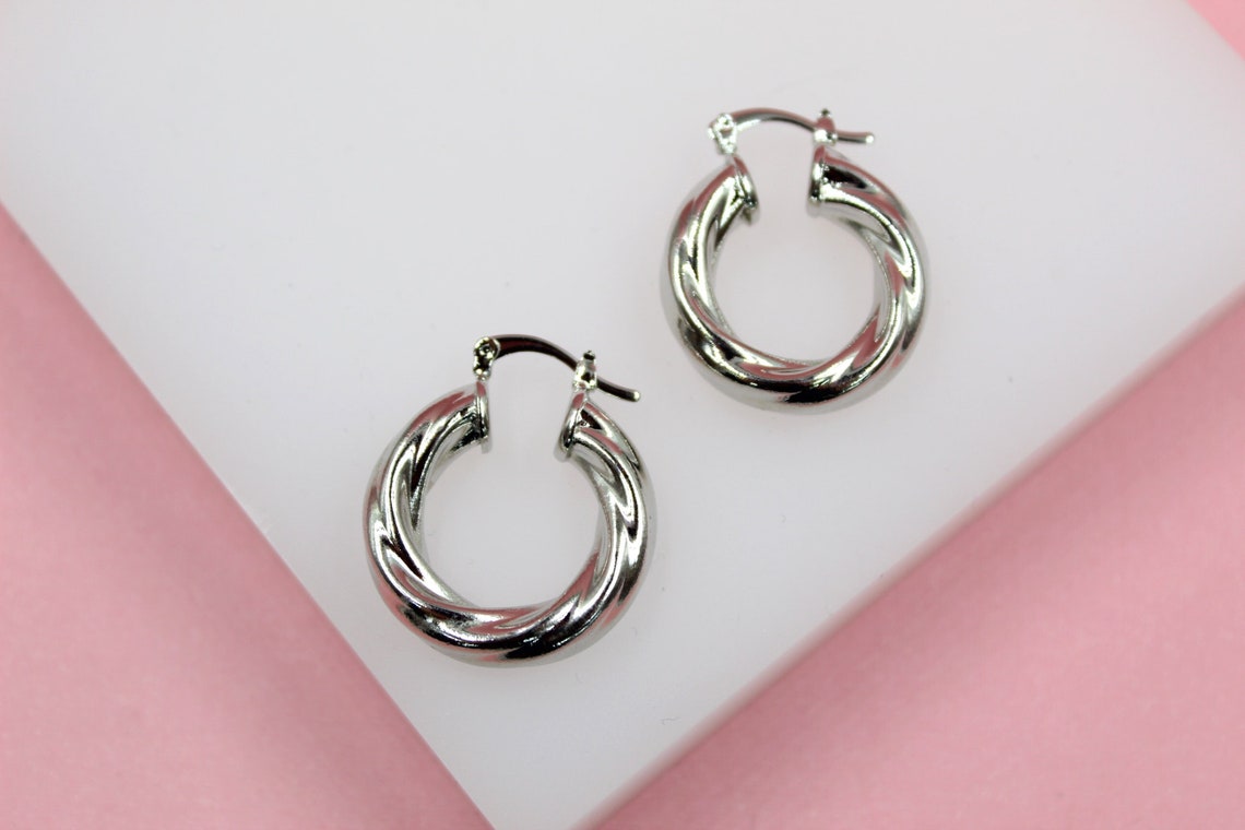 Two polished silver twisted hoop earrings rest on a white display against a soft pink background.