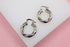 Two polished silver twisted hoop earrings rest on a white display against a soft pink background.