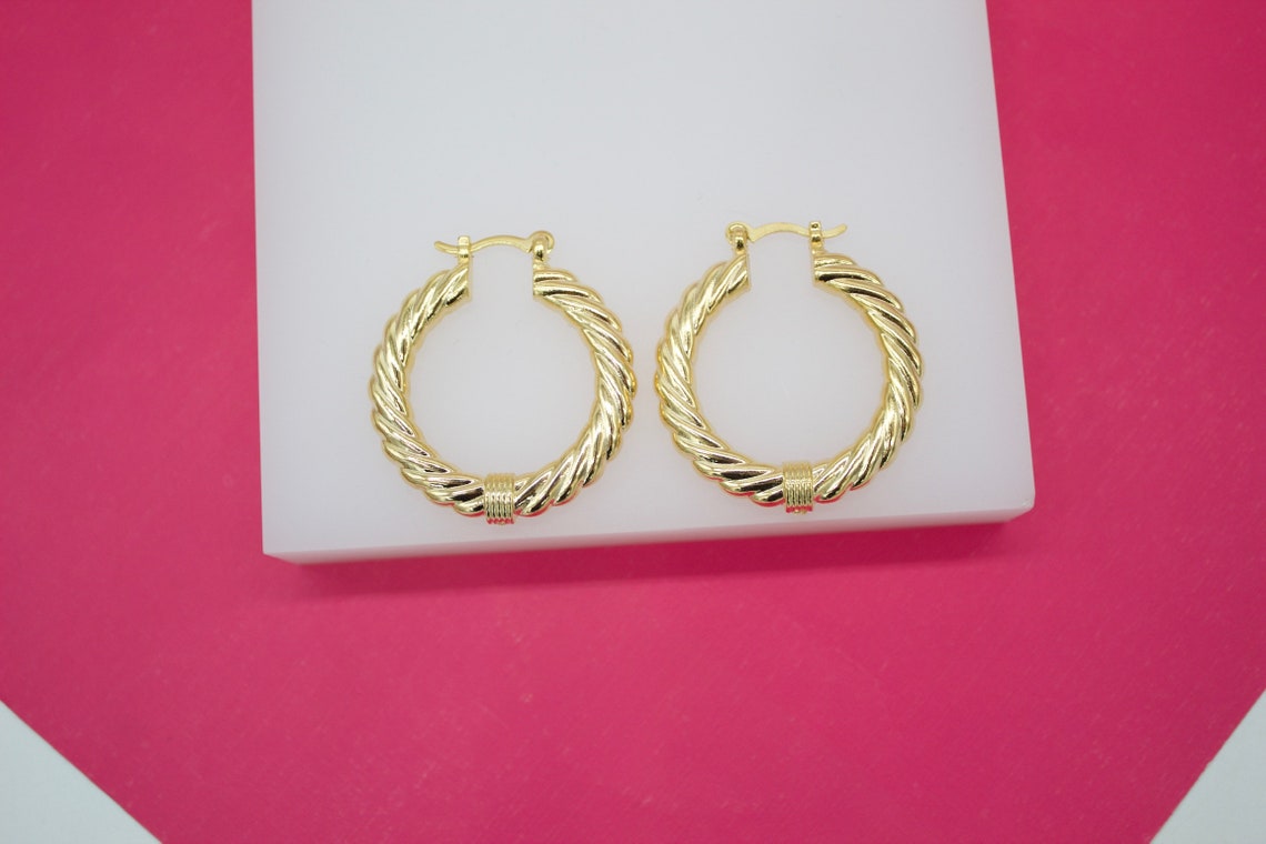 A pair of chunky gold twisted hoop earrings rests on a white display stand against a vibrant pink background.