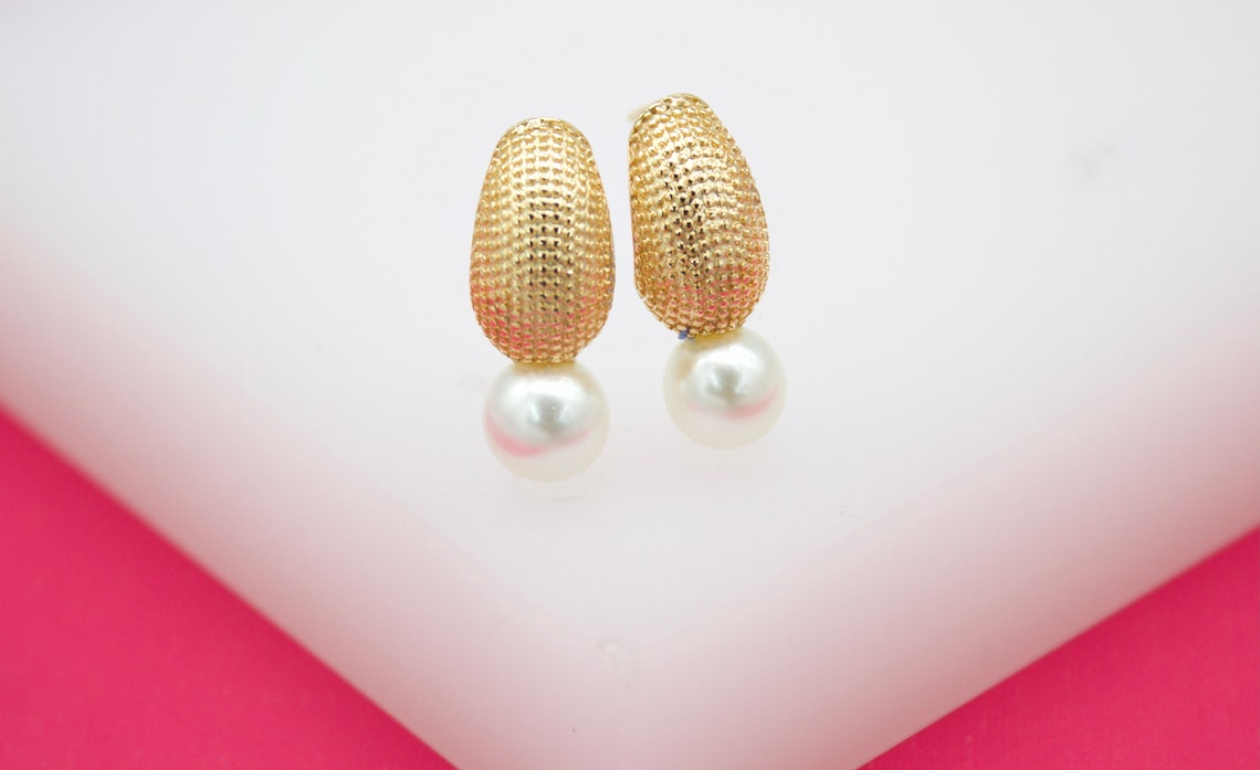 A pair of elegant gold textured drop earrings featuring lustrous white pearls rests on a white surface.