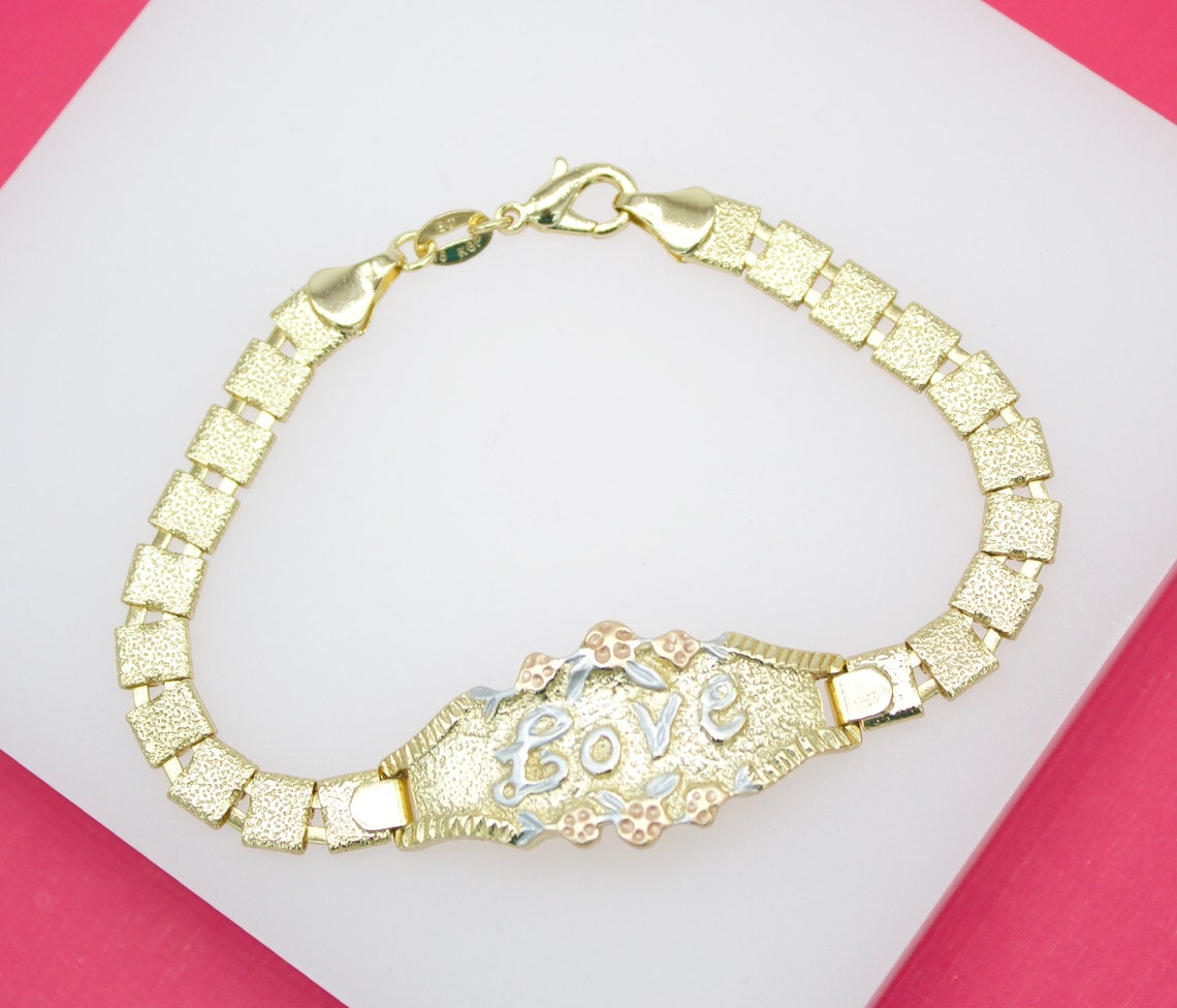 A gold link bracelet featuring an engraved plaque that spells out the word "love" rests on a white display stand against a vibrant pink background.