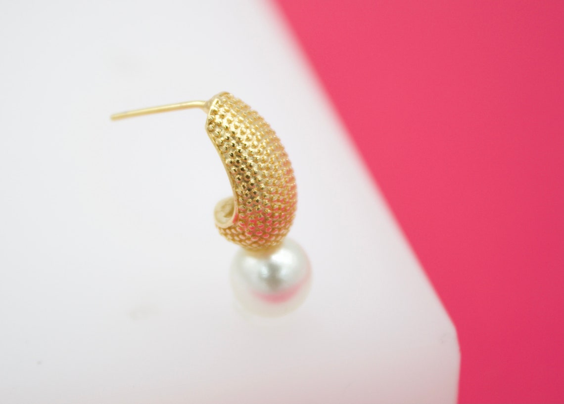 A textured gold hoop earring features a hanging white pearl against a white and pink background.
