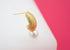 A textured gold hoop earring features a hanging white pearl against a white and pink background.