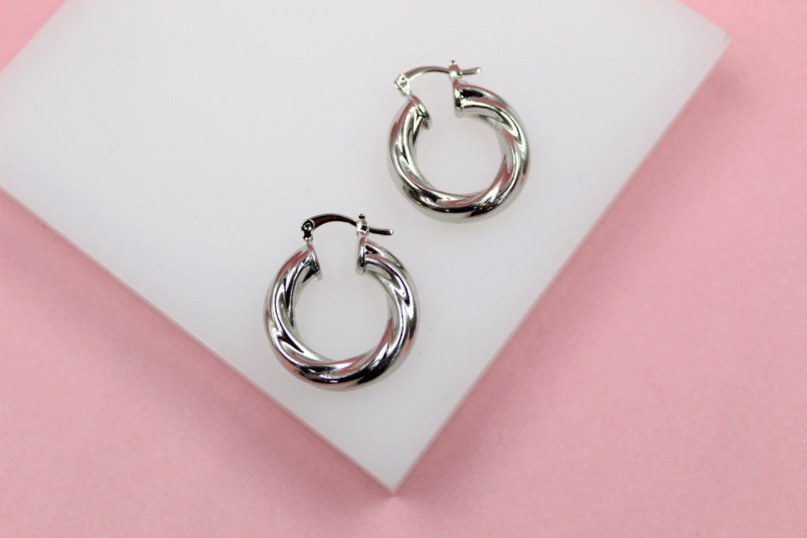A pair of polished silver twist hoop earrings rest on a white display stand against a soft pink background.