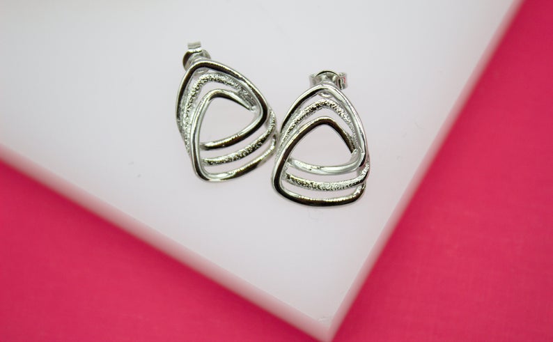 A pair of modern silver geometric stud earrings with textured triangular loops rests on a white display surface against a vibrant pink background.