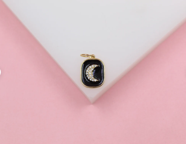 A delicate gold pendant features a sparkling crystal crescent moon set against black enamel on a pink and white background.