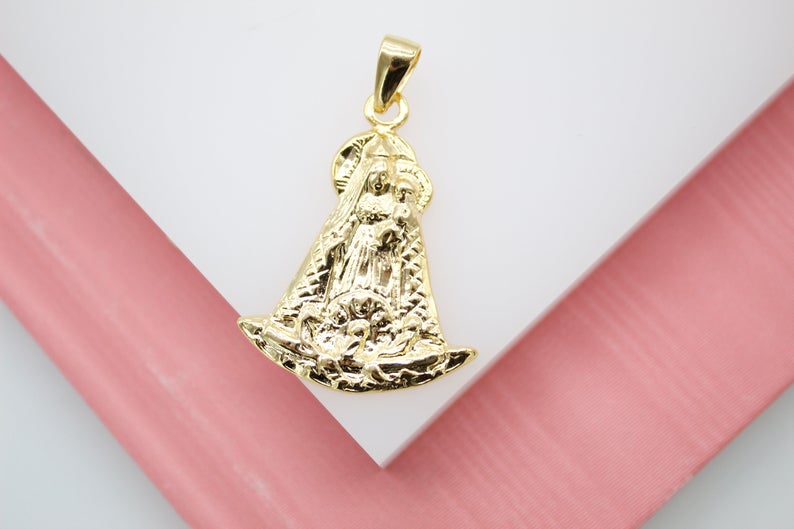 A detailed gold pendant depicting Our Lady of Aparecida rests against a soft pink and white background.