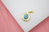 A gold evil eye pendant rests on a white display surface against a pink background.