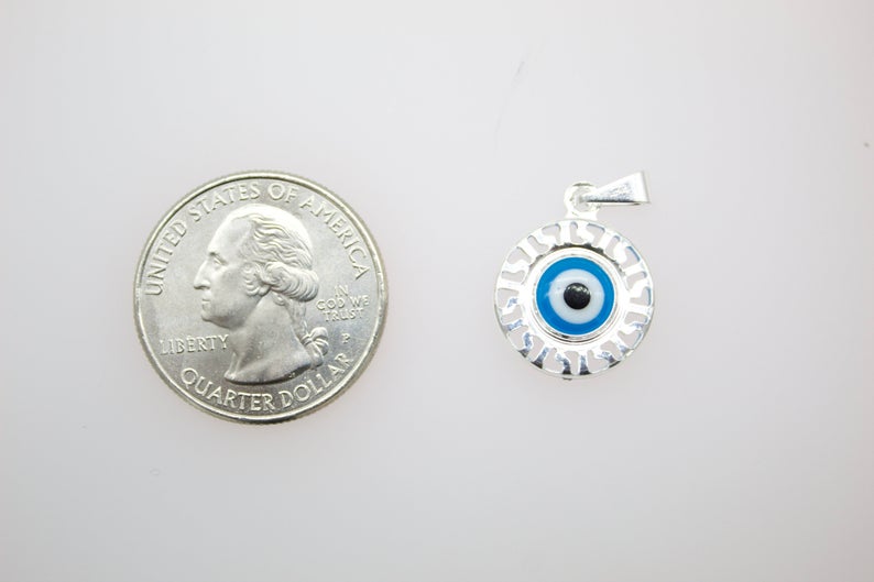 A silver evil eye pendant rests beside a US quarter coin for scale on a light background.