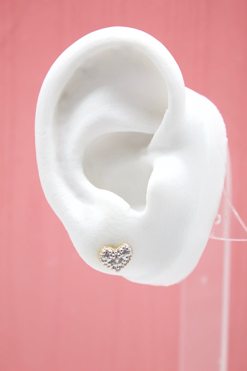 A white mannequin ear displays a sparkling pave diamond heart stud earring against a pink background.