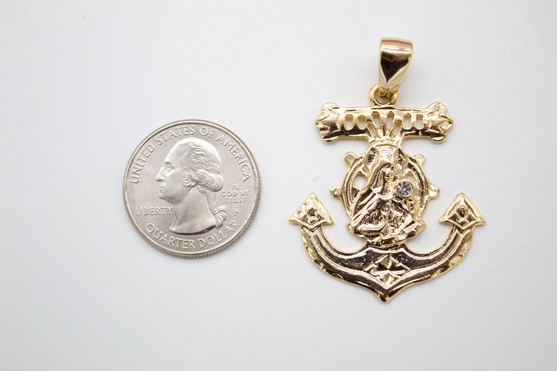 A gold anchor pendant featuring a religious figure at the center rests next to a US quarter coin for scale.