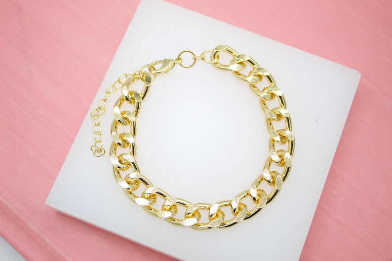 A thick gold chain link bracelet rests neatly on a white display block against a soft pink background.