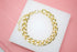 A thick gold chain link bracelet rests neatly on a white display block against a soft pink background.