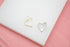 Two heart-shaped druzy pendants, one gold and one silver, rest on a white surface against a pink background.