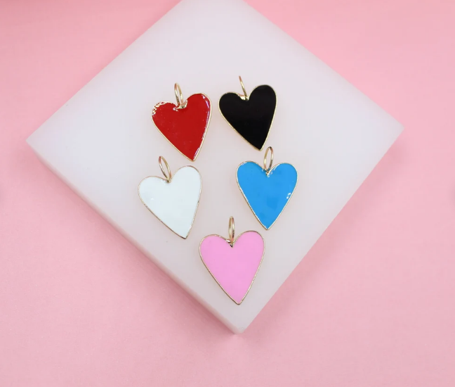Five colorful heart-shaped enamel charms with gold trim rest on a white display block against a pink background.