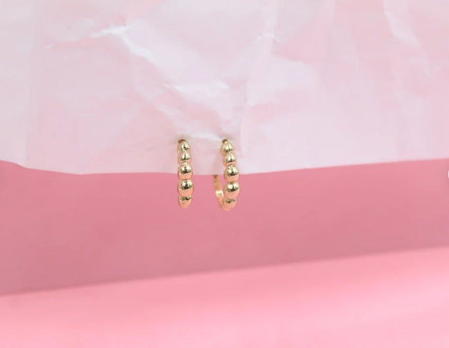 A pair of gold beaded huggie hoop earrings rests against a pink and white textured backdrop.