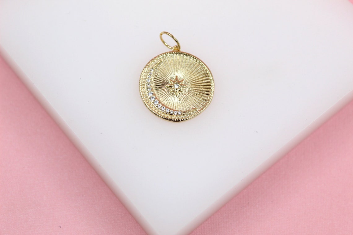 A round gold celestial pendant featuring a sparkling diamond crescent moon and star rests on a white display surface against a pink background.