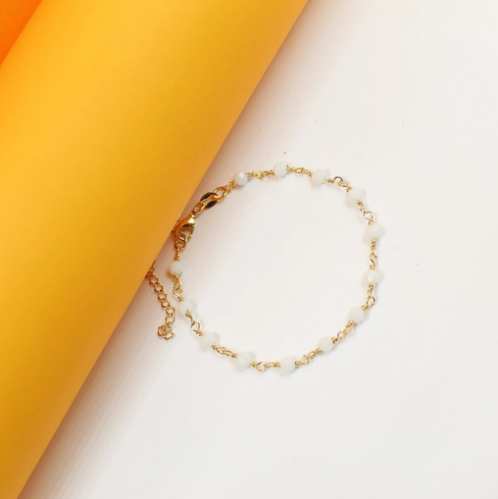A delicate gold chain bracelet features small white beads arranged symmetrically against a bright yellow and white background.