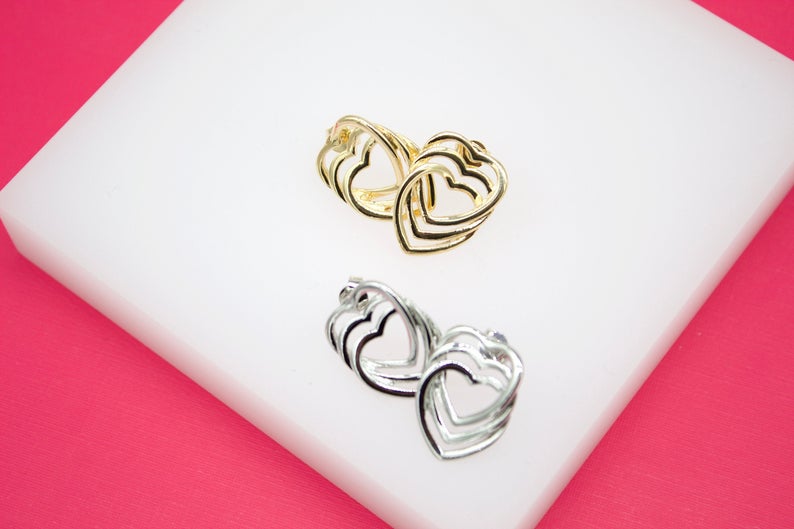 A pair of gold and silver heart-shaped hoop earrings rests on a white display surface against a bright pink background.