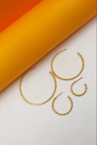 Four gold twist hoop earrings of varying sizes rest on a white surface against an orange and yellow curved background.
