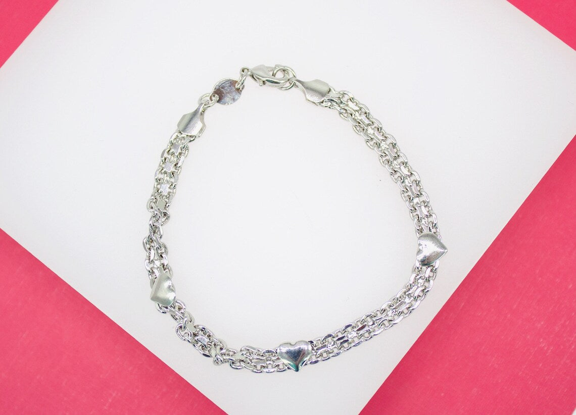 A delicate silver double-strand charm bracelet rests on a white and pink background, featuring intricate link chains adorned with small heart charms.