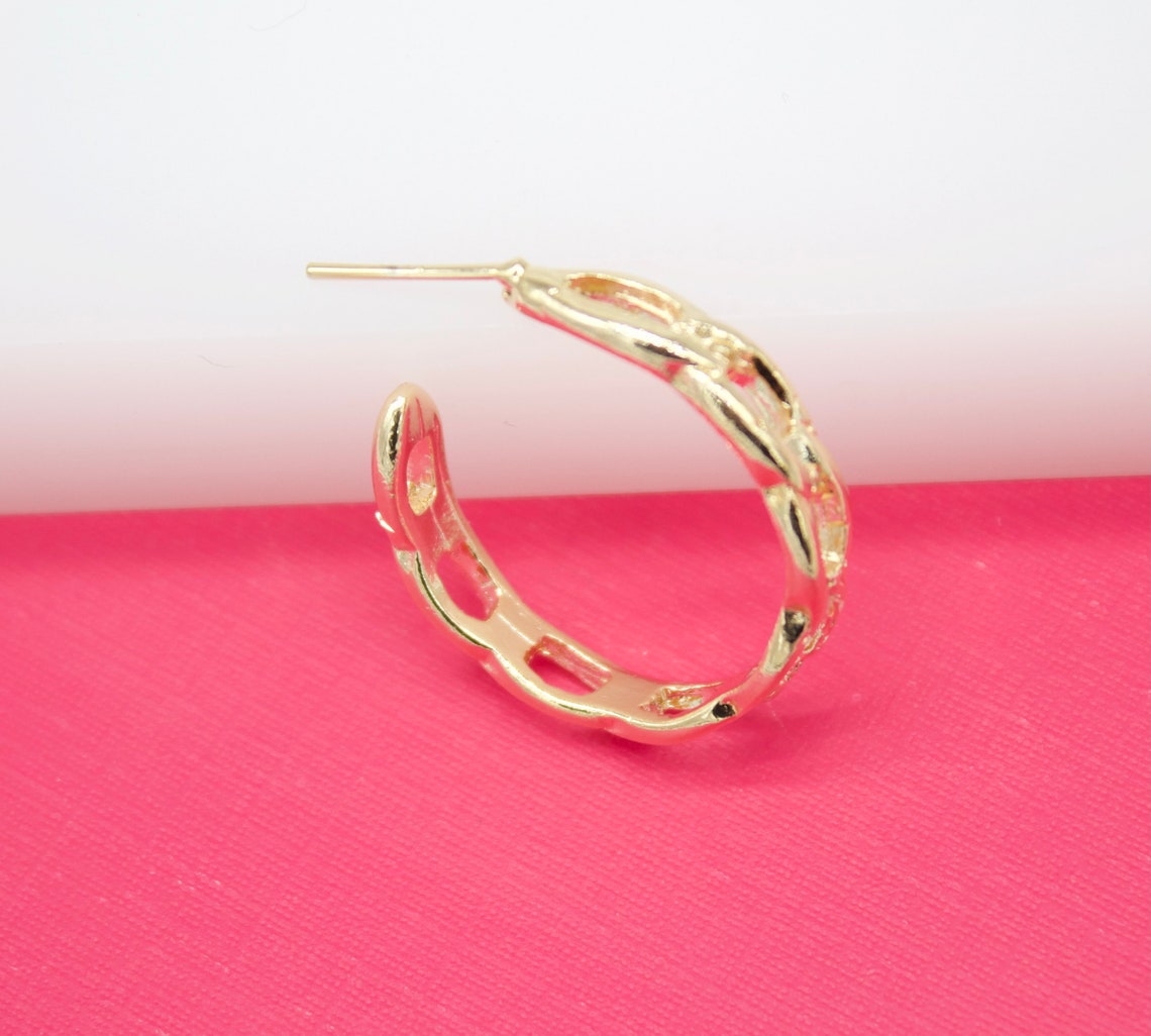 A polished gold hoop earring with a cutout link pattern rests on a vibrant pink surface.