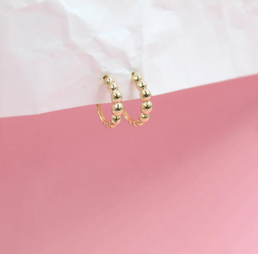 A pair of gold beaded hoop earrings is displayed against a white and pink background.