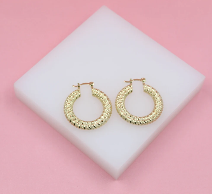 A pair of textured gold hoop earrings rests on a white display block against a pink background.