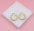 A pair of textured gold hoop earrings rests on a white display block against a pink background.