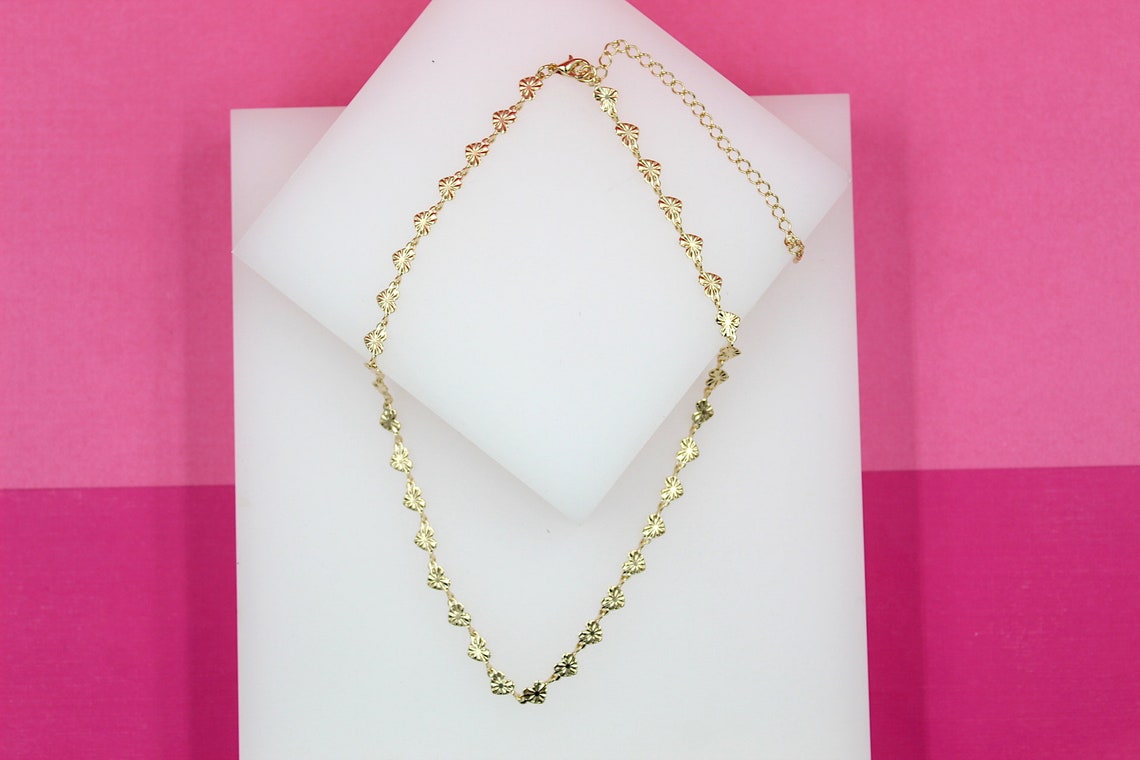 A delicate gold-plated heart-link chain necklace rests elegantly on white display blocks against a vibrant pink background.