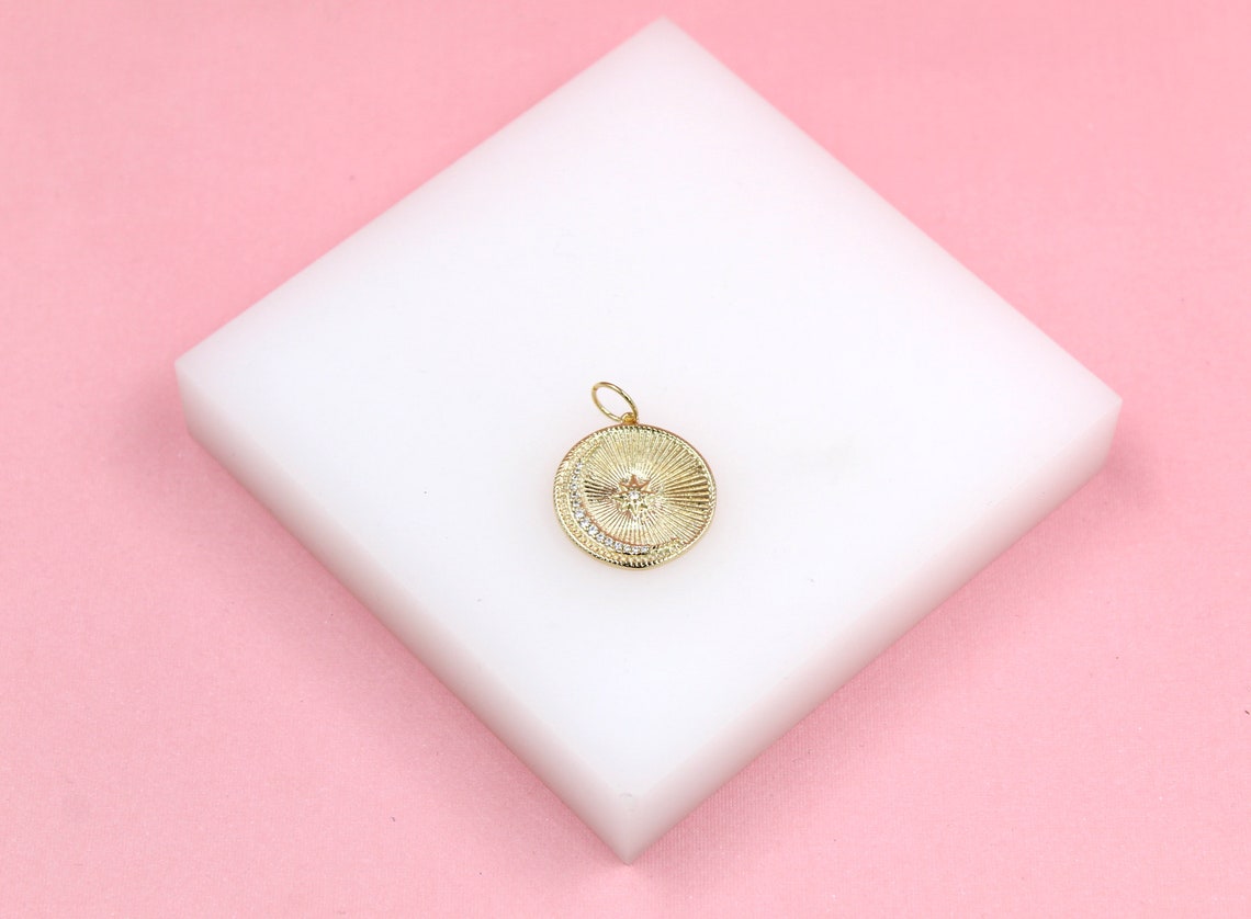 A gold celestial pendant featuring a sunburst design with a crystal crescent moon rests on a white block against a pink background.