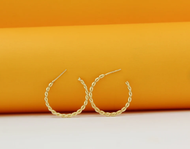 A pair of gold twisted hoop earrings rests against a vibrant orange and white background.
