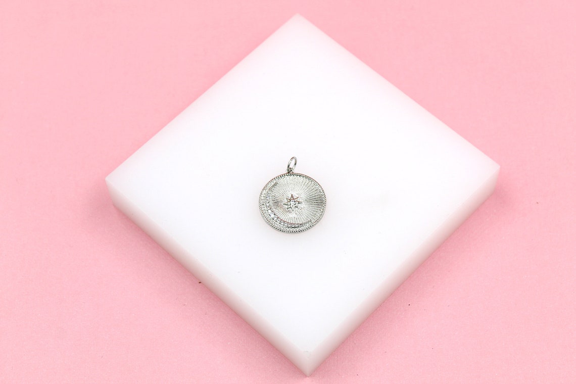 A silver celestial pendant with a crescent moon and star design rests on a white display block against a pink background.