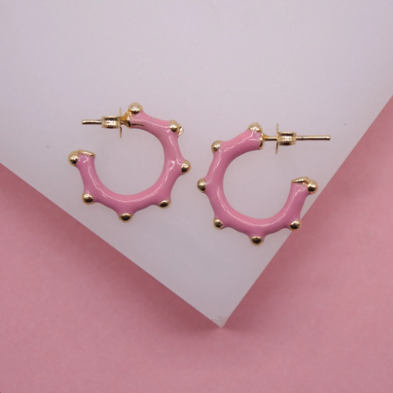 A pair of pink enamel hoop earrings with gold-plated studs rest on a white display surface against a pink background.