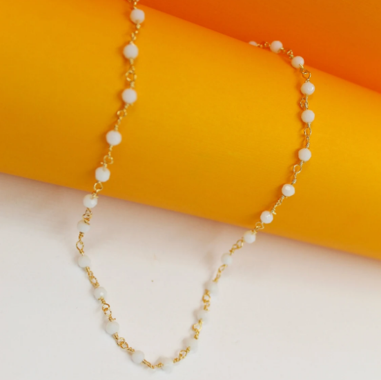 A delicate gold chain necklace with evenly spaced white beads drapes gracefully across a bright orange and white backdrop.