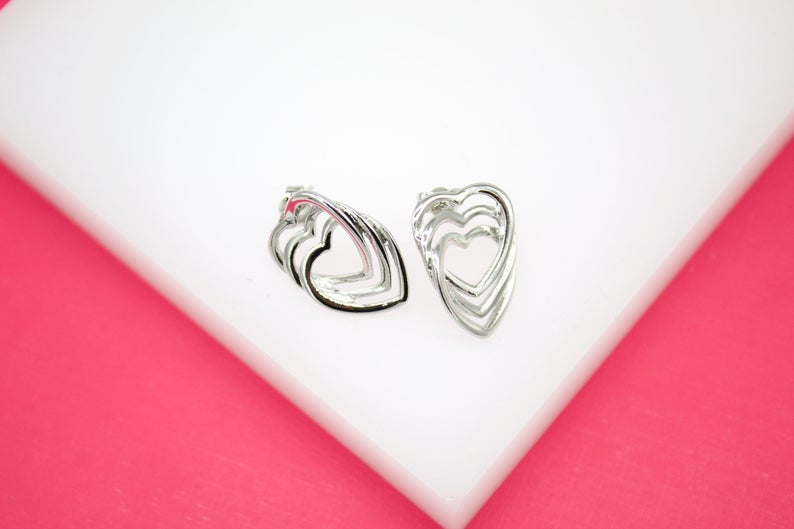 A pair of silver multi-heart stud earrings rests on a white display surface against a pink background.