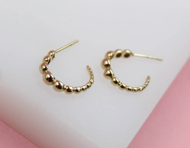 A pair of gold beaded hoop earrings rests on a white display surface.