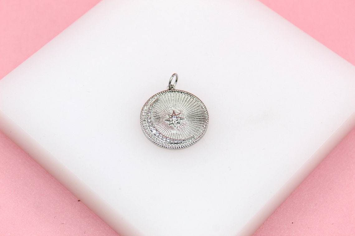 A silver celestial coin pendant featuring a crescent moon and a sparkling star rests on a white display against a pink background.